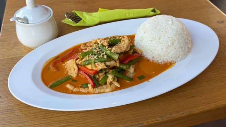 Pa Naeng Curry mit Hähnchen, grünen Bohnen, Paprika und Erdnüssen neben Jasminreis auf weißem Teller